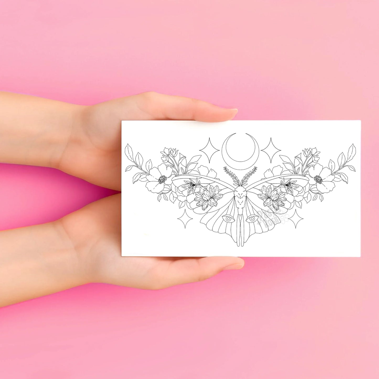 Hands holding a temporary tattoo with a black floral and moon design on a pink background