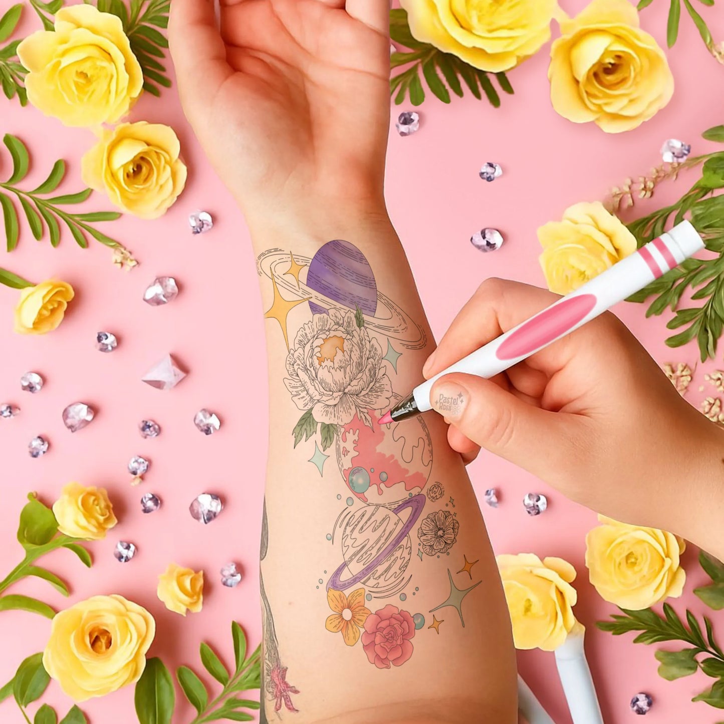 Hand with temporary tattoo design on arm, surrounded by yellow flowers and decorative elements on a pink background.
