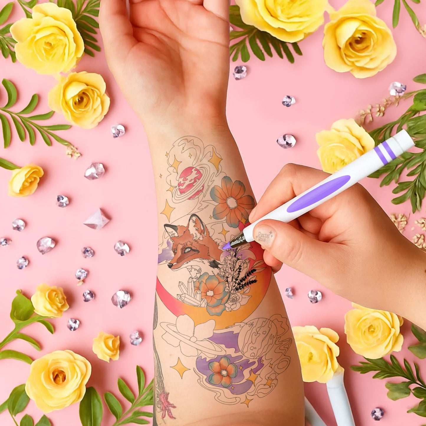 Hand applying a temporary tattoo of a fox and flowers on a pink background with yellow roses and decorative elements.