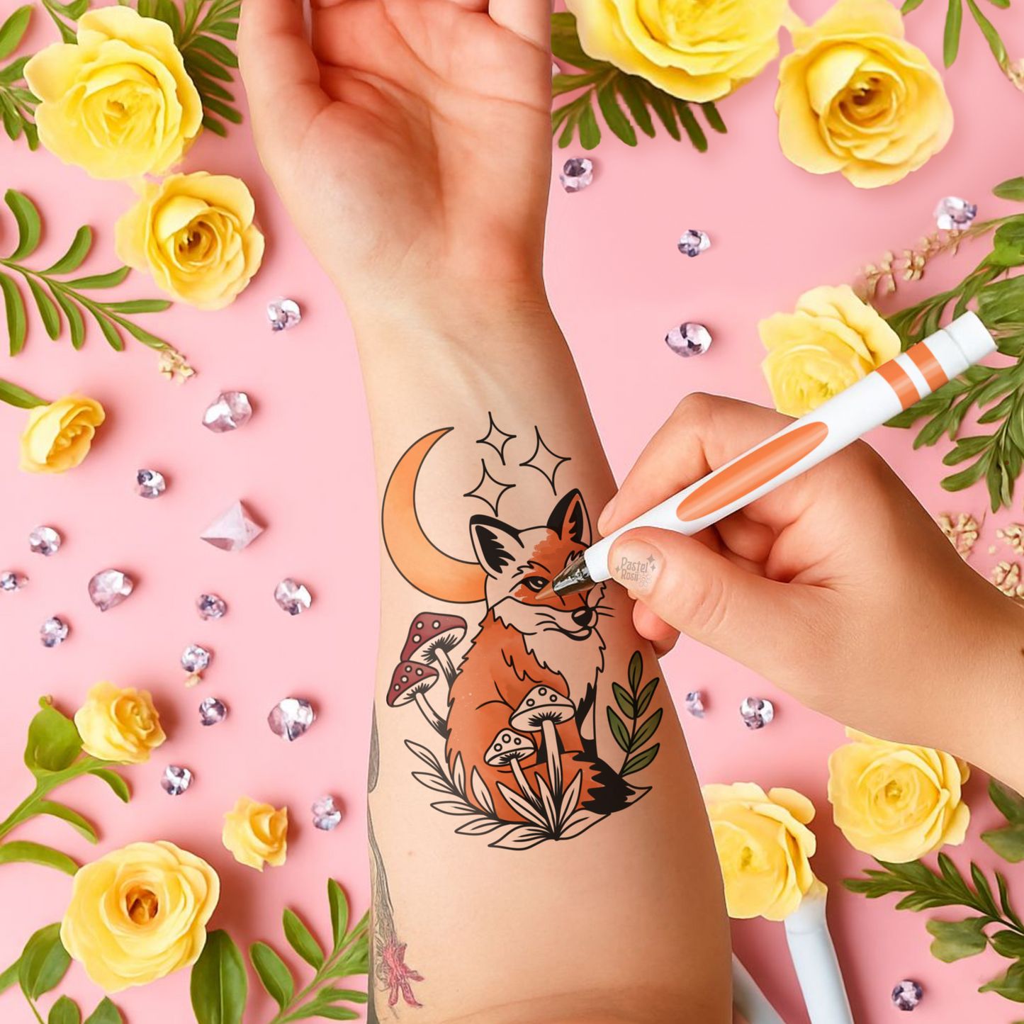 Hand applying a temporary tattoo of a fox and mushrooms design on a pink background with yellow flowers and green leaves.