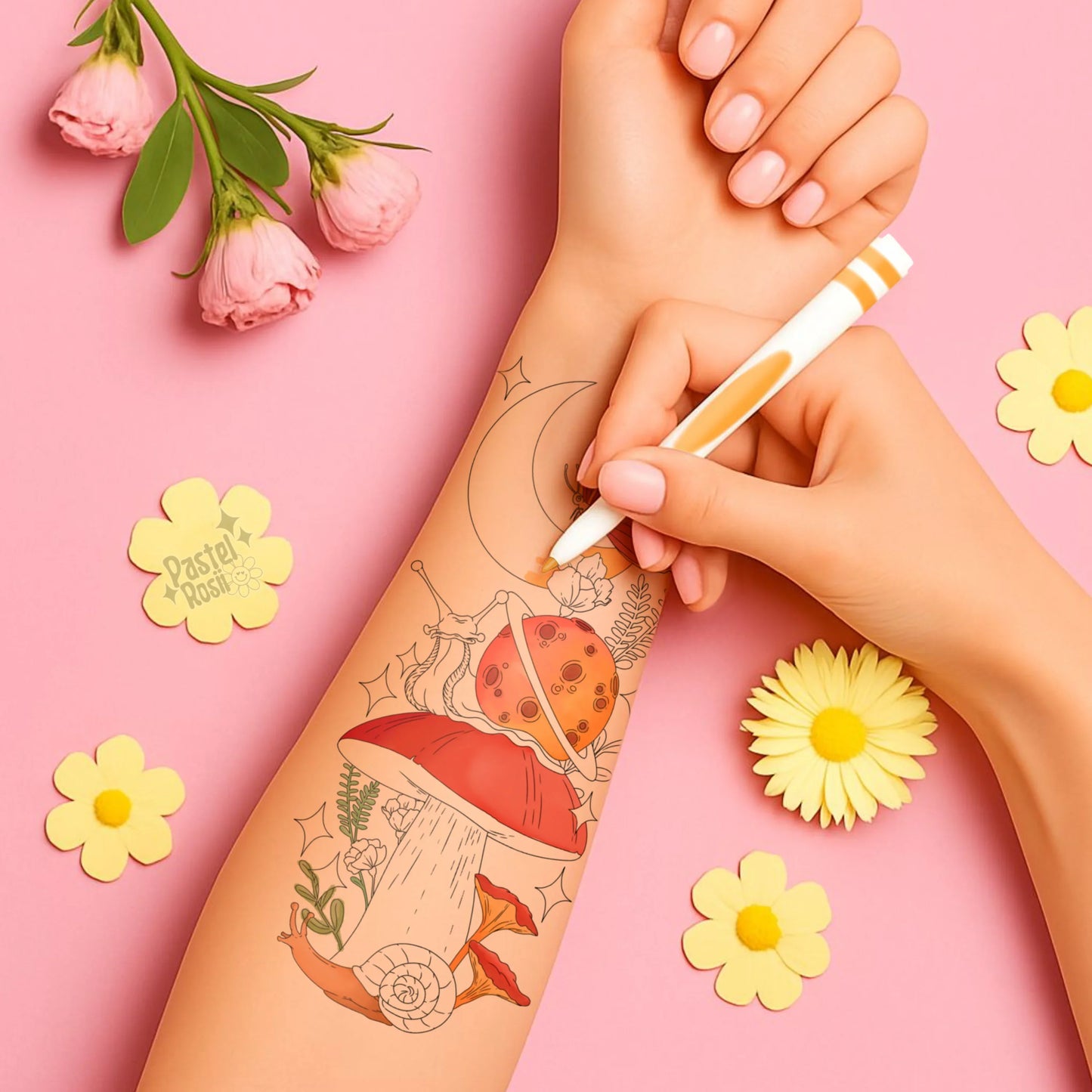 A hand draws on a tattoo design featuring a mushroom, planets, and flowers on a pink background, surrounded by fresh blooms.