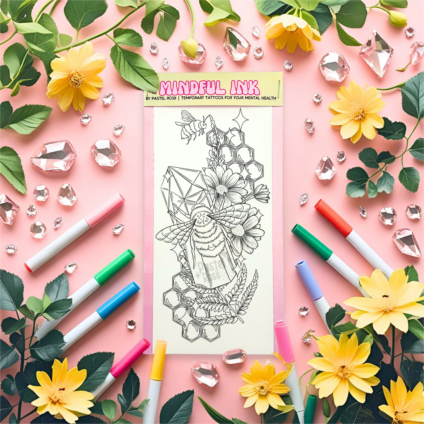 A black-and-white temporary tattoo design featuring a bee, flowers, and crystals, surrounded by colorful markers and decorative gems.