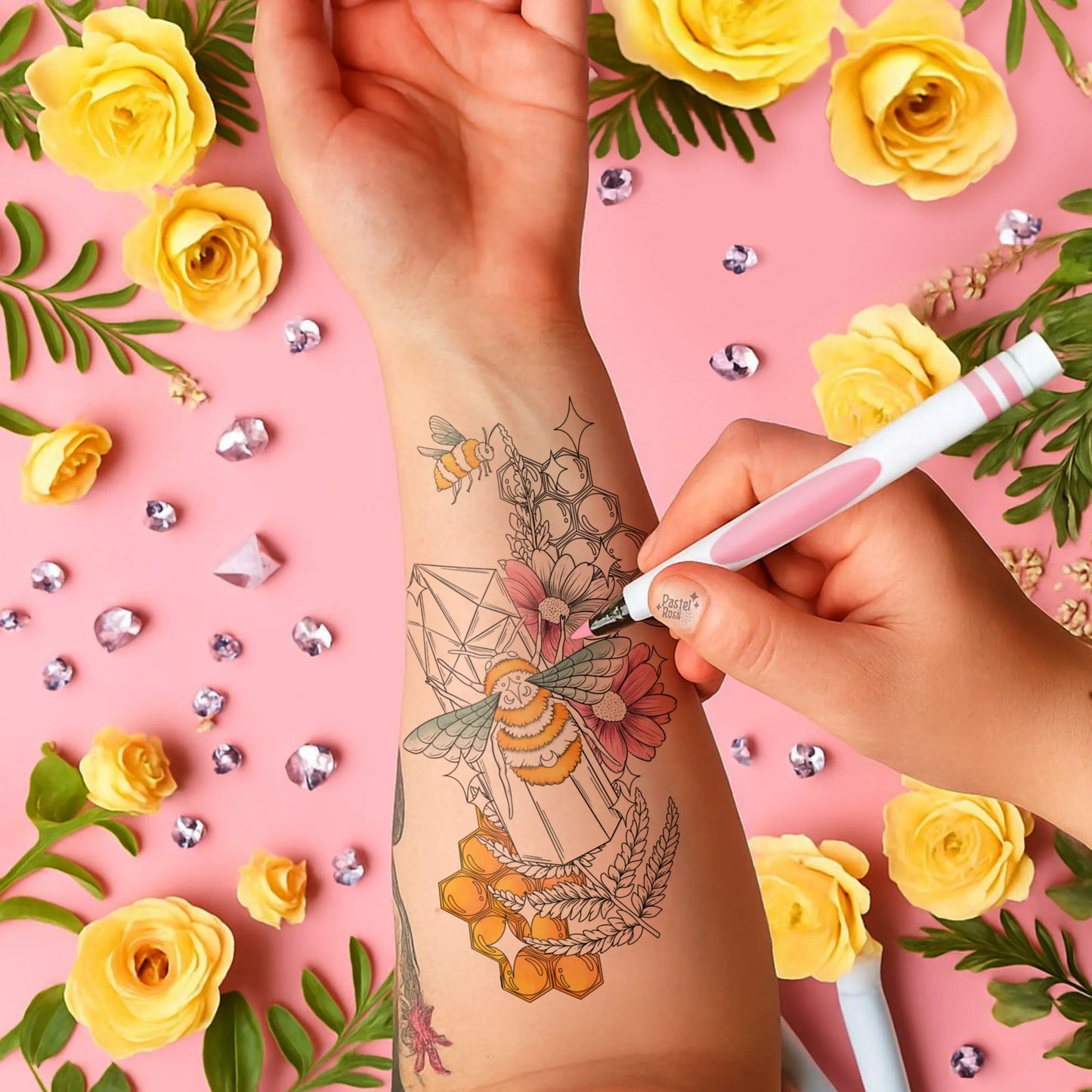 A hand colors a tattoo of bees and flowers on an arm, surrounded by yellow roses and pink background accents.