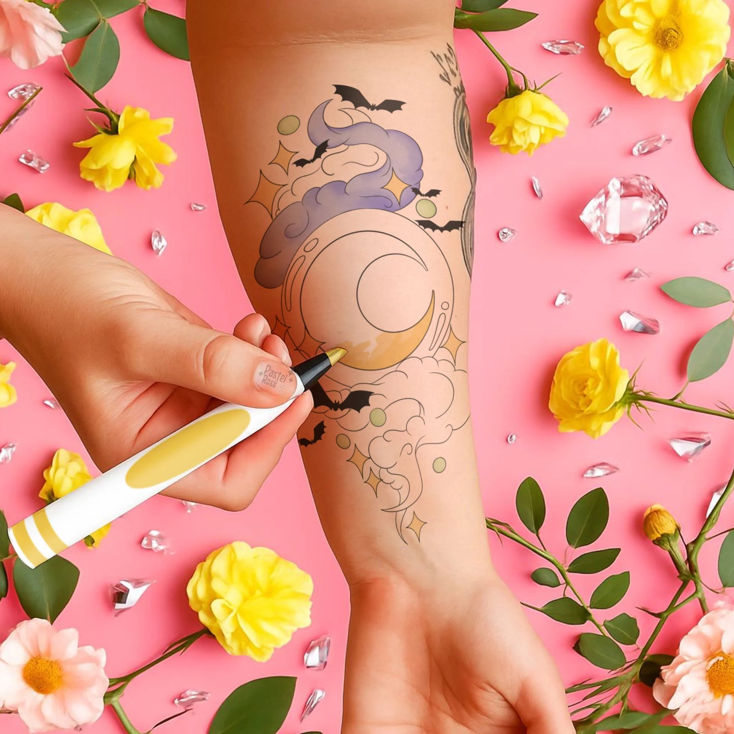 Person applying temporary tattoo of a witch with a moon on a pink background with flowers and crystals.