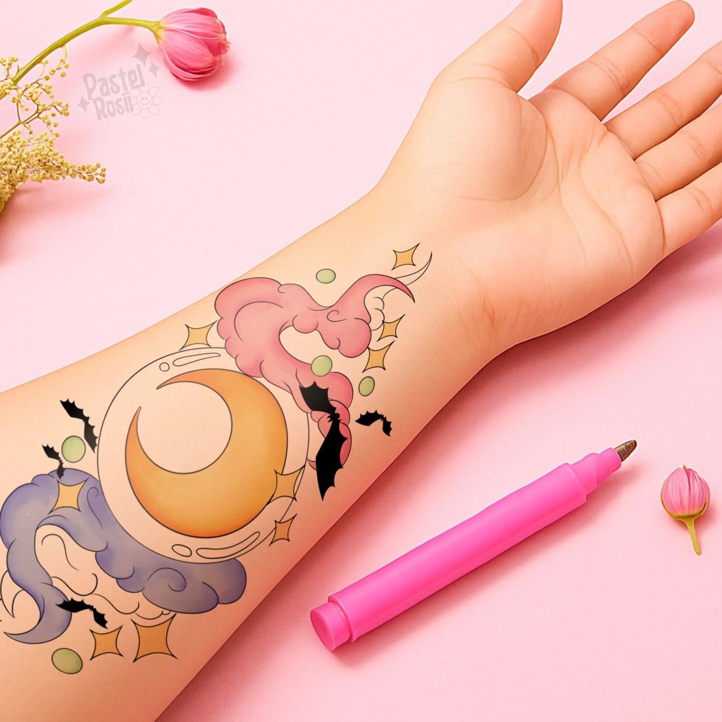 Hand with temporary tattoo of a moon and cloud design on a pink background with a pink pen.