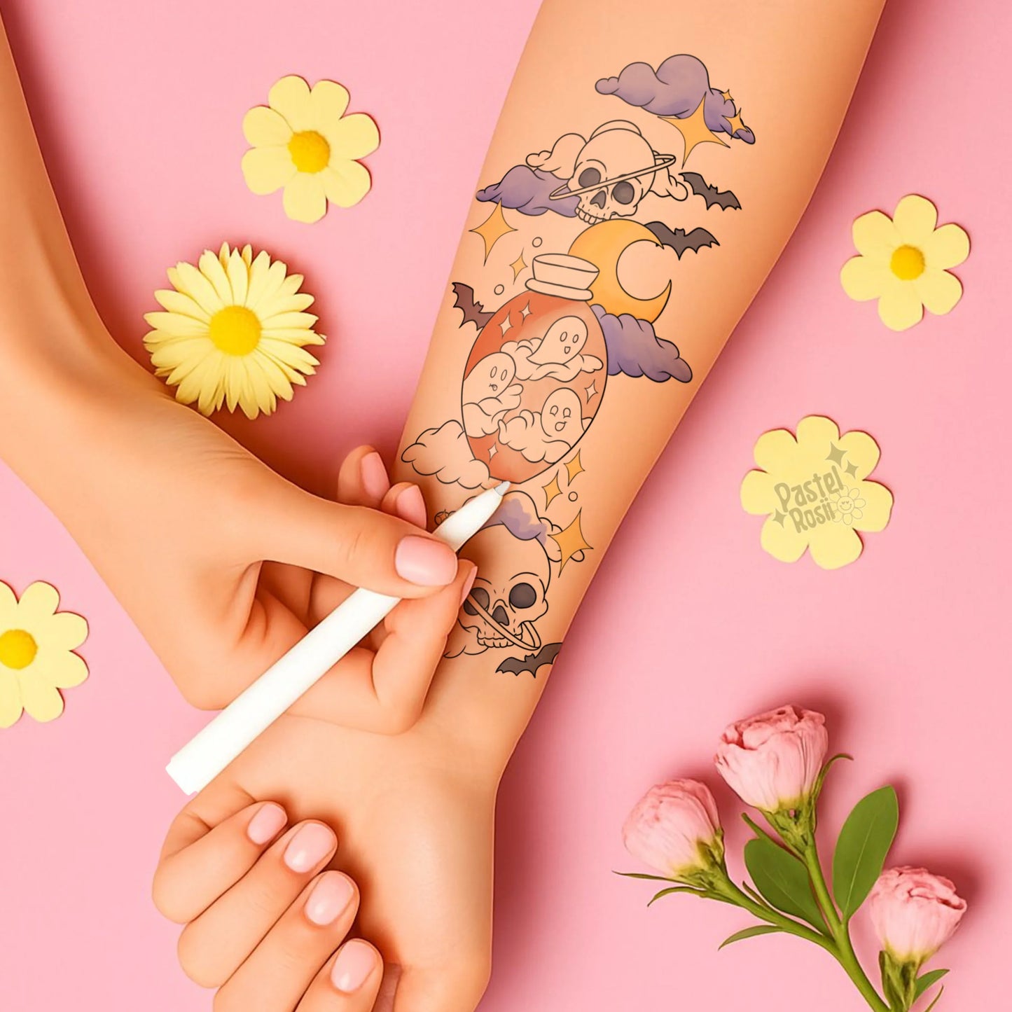 Hand applying temporary tattoo with skull and moon design on pink background with flowers