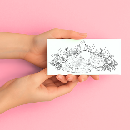 Hands holding a temporary tattoo with a black and white illustration of a cat, crystals, and flowers on a pink background.