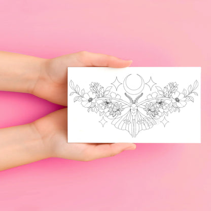 Hands holding a temporary tattoo with a black floral and moon design on a pink background