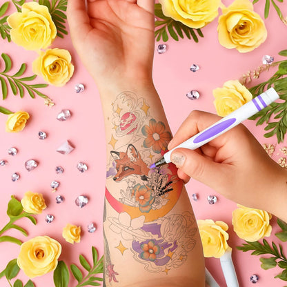 Hand applying a temporary tattoo of a fox and flowers on a pink background with yellow roses and decorative elements.