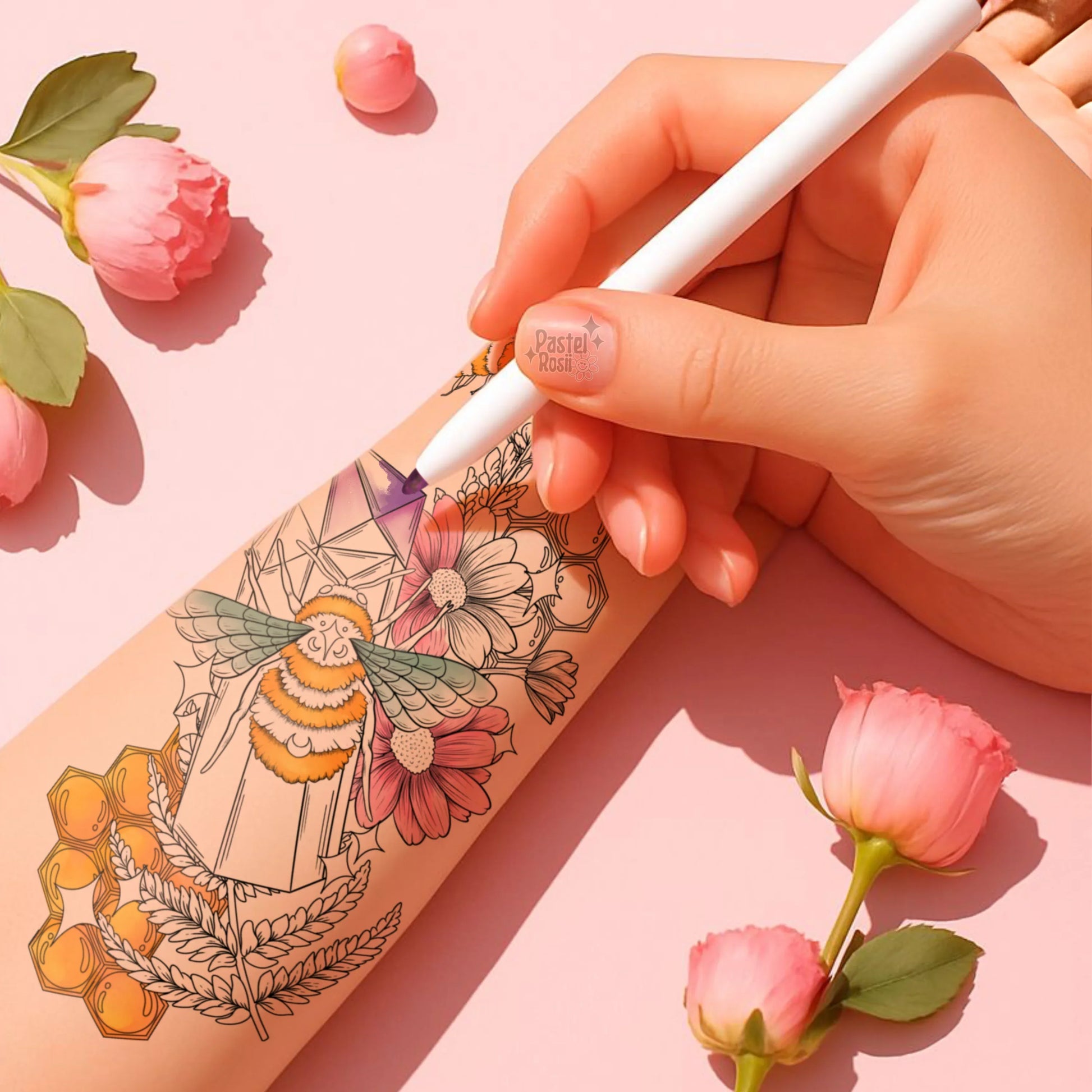 Hand holding a white pen coloring a temporary tattoo on a pink background with flowers