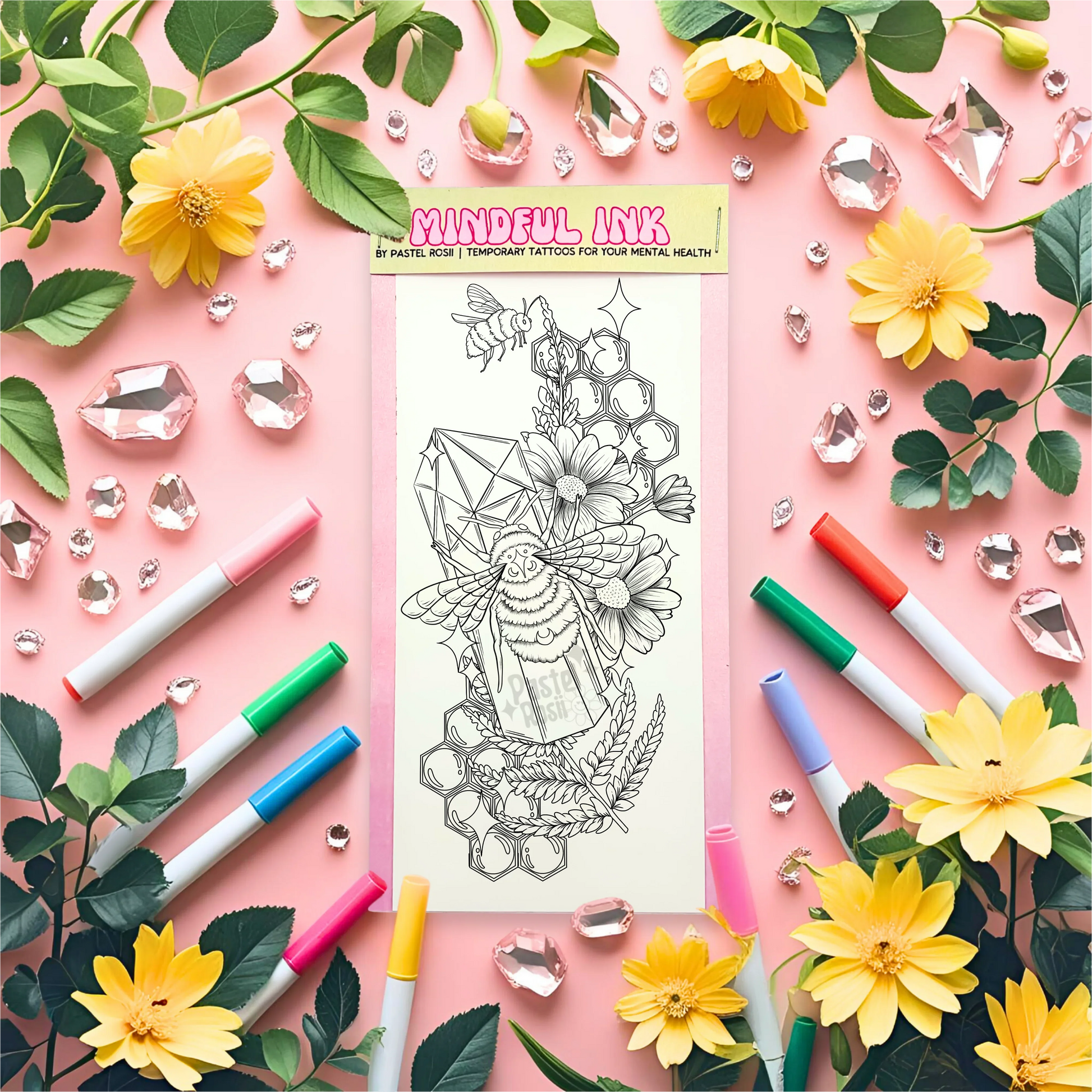 A black-and-white temporary tattoo design featuring a bee, flowers, and crystals, surrounded by colorful markers and decorative gems.